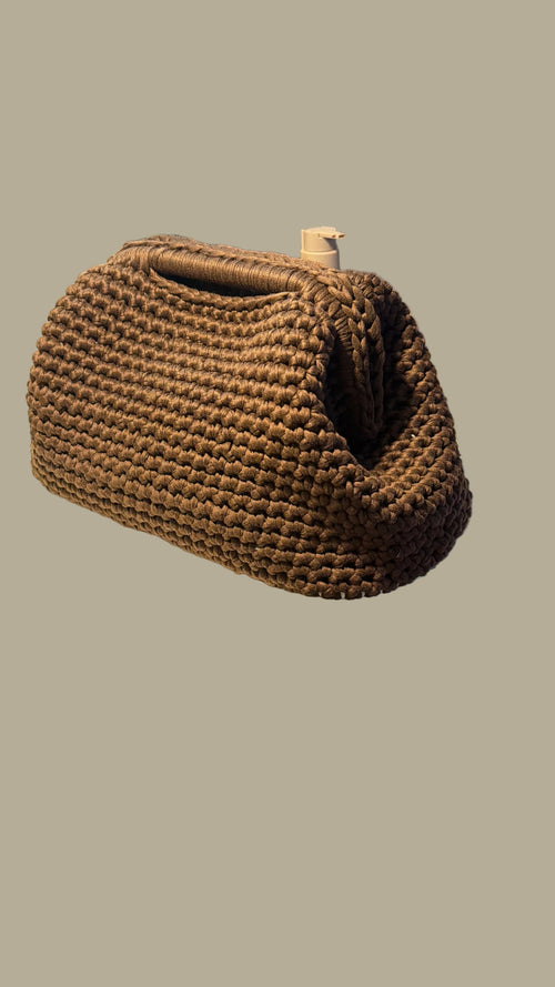A brown woven basket-style clutch bag resting on a white vanity shelf in a real interior setting, with decorative greenery and a mirror visible.